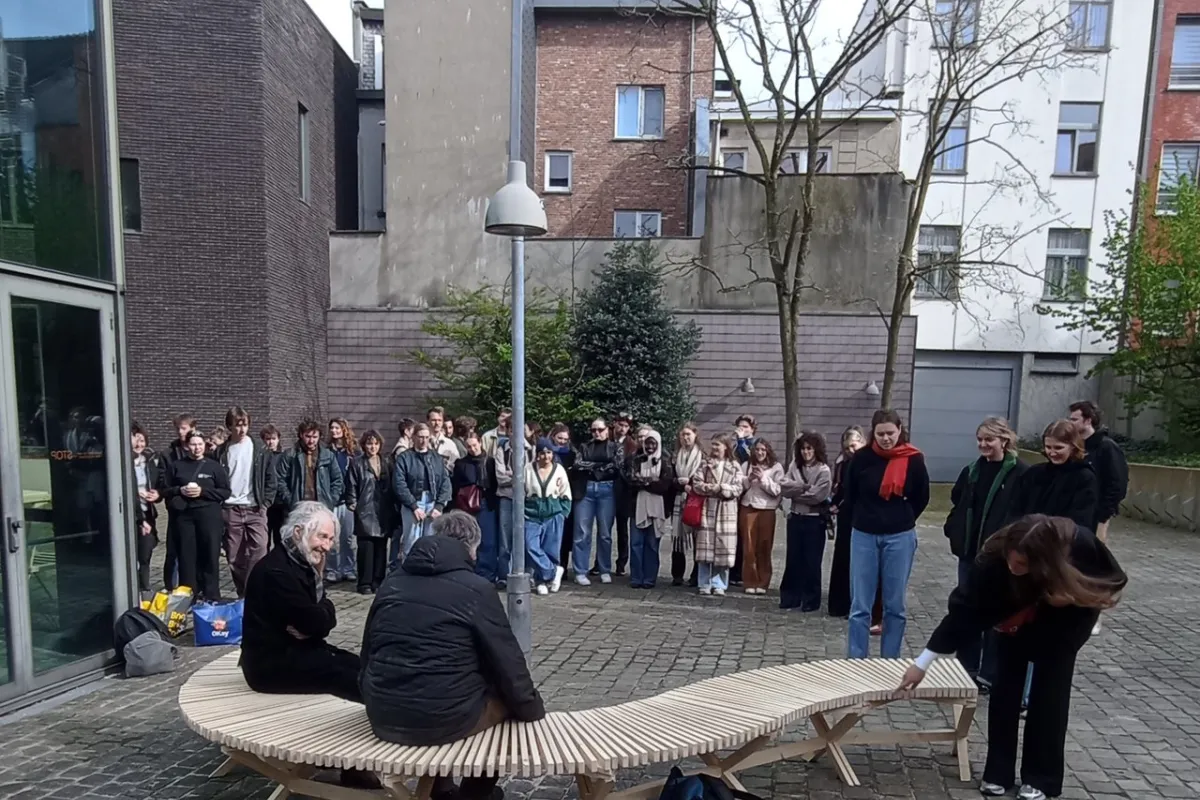 Eine Gruppe von Studierenden steht im Freien um eine geschwungene Holzbank, die von zwei Personen genutzt wird. Die Szene findet auf einem gepflasterten Platz vor modernen Gebäuden statt.