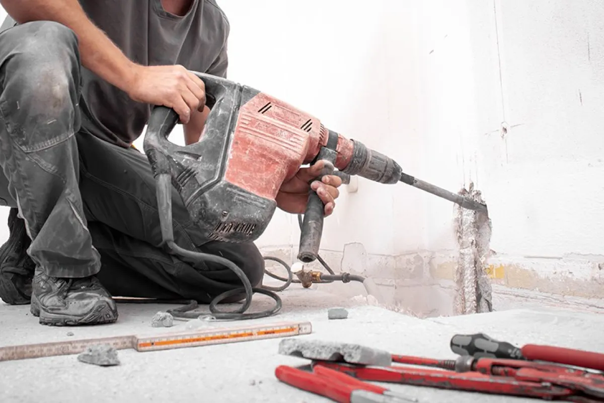 Ein Bauarbeiter kniet auf dem Boden und benutzt einen roten Kombi-Hammer, um eine Wand zu bearbeiten. Im Vordergrund liegen Werkzeuge und Bauutensilien.