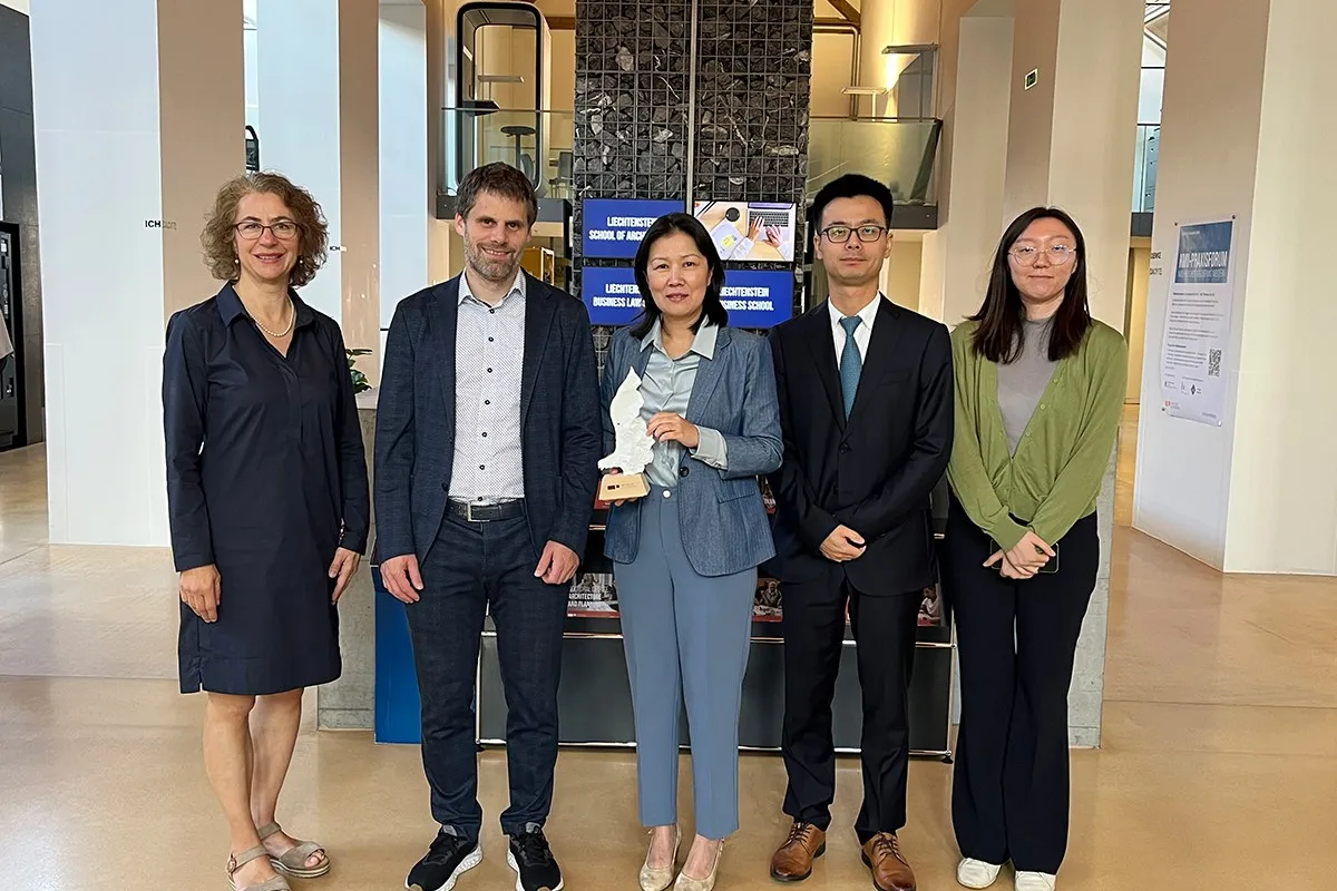 Eine Gruppe von fünf Personen steht in einem modernen Gebäude der Universität Liechtenstein. Sie lächeln in die Kamera. Im Hintergrund sind Informationsschilder der Universität zu sehen.