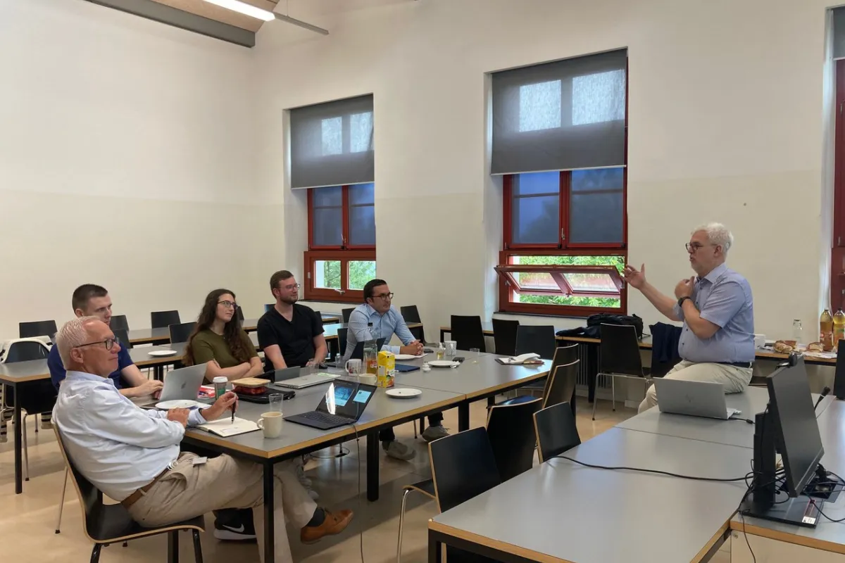 Ein Dozent hält einen Vortrag vor einer kleinen Gruppe von Personen in einem Seminarraum. Die Teilnehmenden sitzen an Tischen mit Laptops und Notizbüchern. Im Hintergrund sind Fenster und eine Leinwand zu sehen.