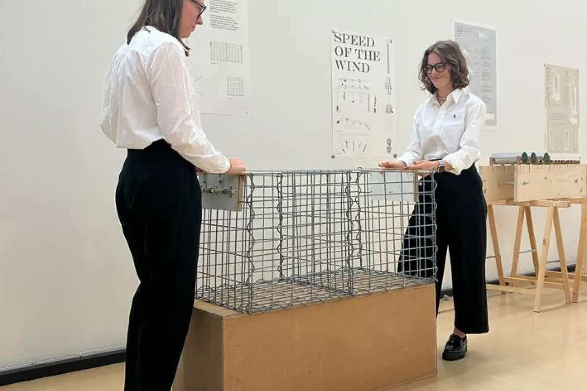 Zwei Personen in weißer Kleidung stehen in einem Ausstellungsraum der Universität Liechtenstein. Sie betrachten ein Modell aus Drahtgeflecht auf einem Holzsockel. Im Hintergrund sind Poster mit Architekturzeichnungen zu sehen.