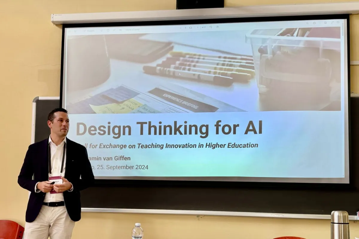 Ein Mann steht vor einer Leinwand, auf der eine Präsentation mit dem Titel "Design Thinking for AI" gezeigt wird. Der Raum wirkt wie ein Seminarraum, und es sind einige Stifte und Notizen auf der Leinwand zu sehen.