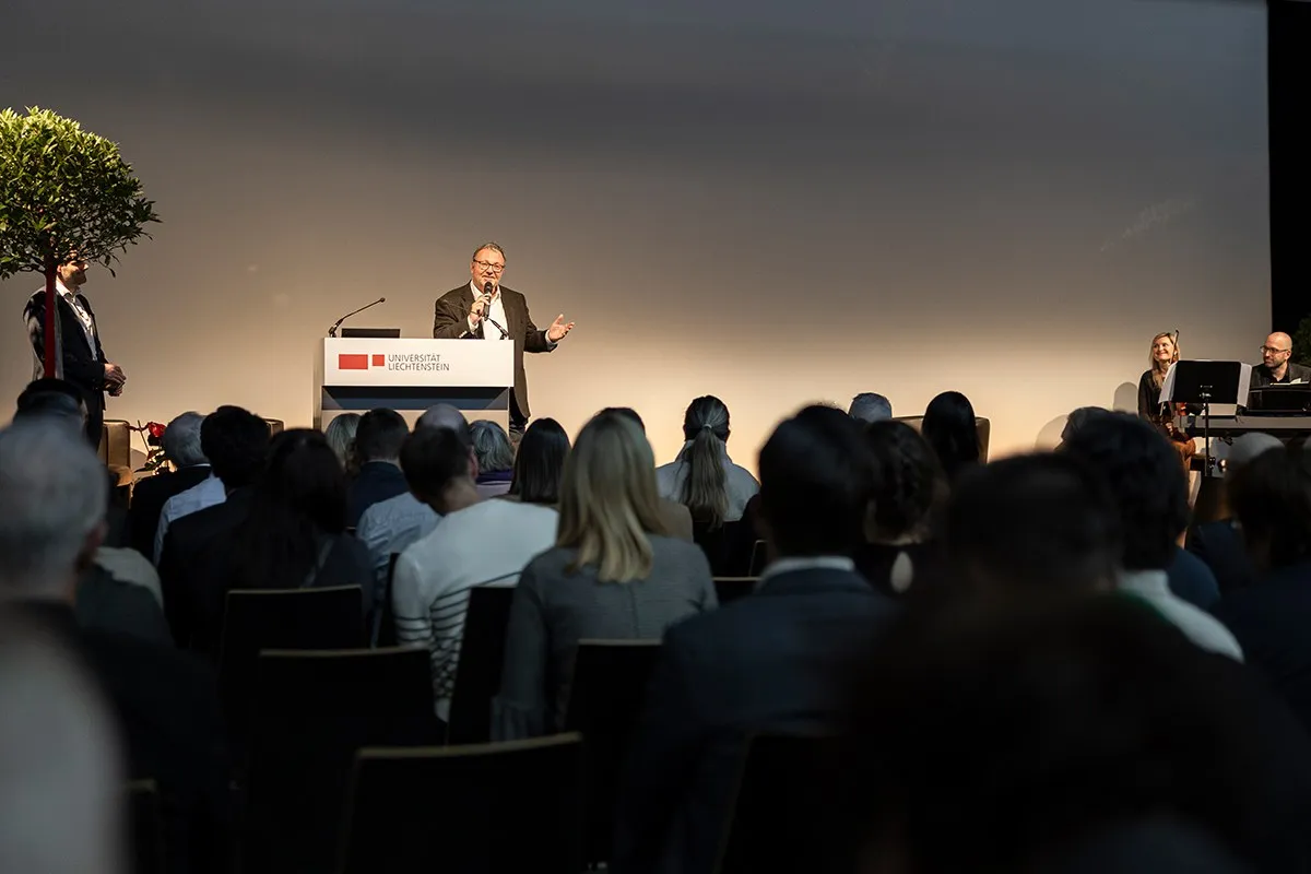 Ein Redner steht an einem Podium mit dem Logo der Universität Liechtenstein, spricht zu einem Publikum. Im Hintergrund sind zwei Musiker zu sehen. Die Veranstaltung findet in einem großen Saal statt.