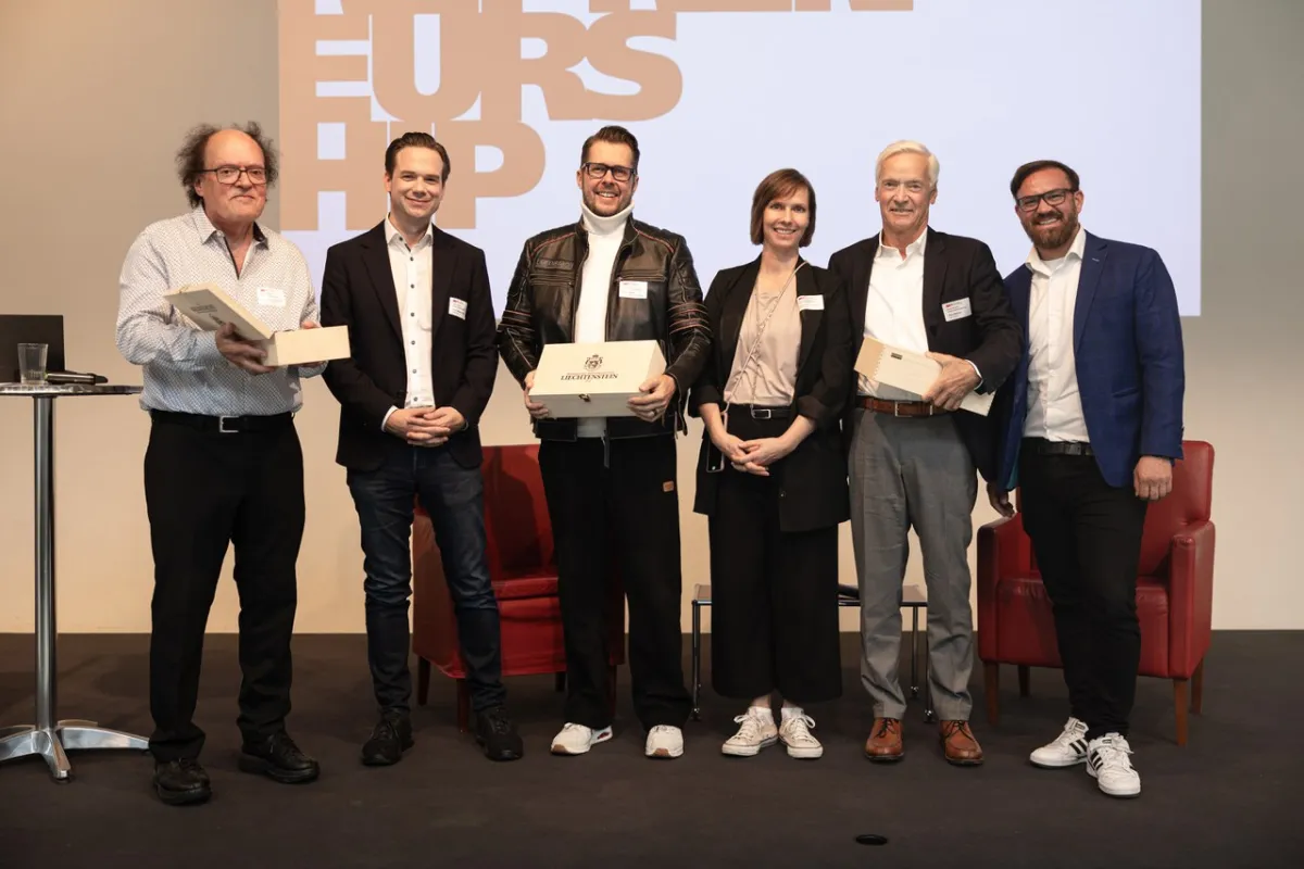 Sechs Personen stehen auf einer Bühne und halten Geschenke in den Händen. Im Hintergrund ist ein Bildschirm mit dem Wort "Entrepreneurship" zu sehen. Die Personen lächeln und tragen Namensschilder.