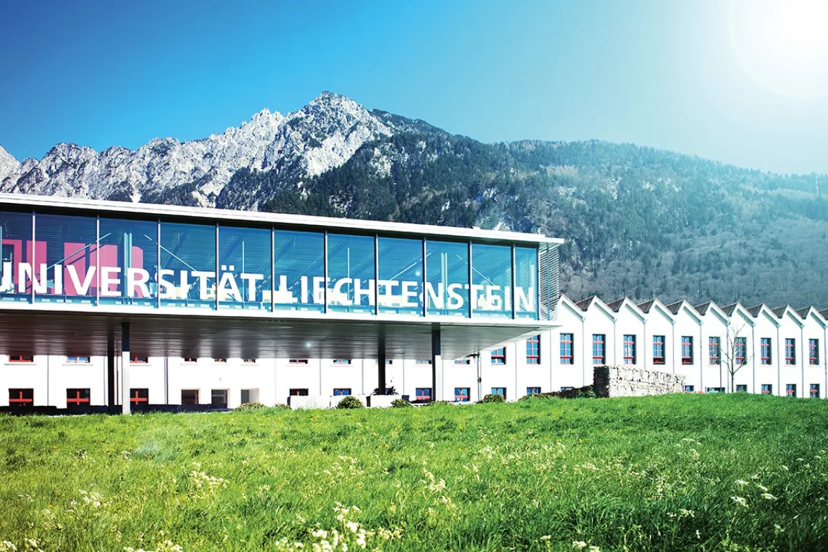 Außenansicht der Universität Liechtenstein mit modernem Glasgebäude vor malerischer Bergkulisse, umgeben von grüner Wiese.