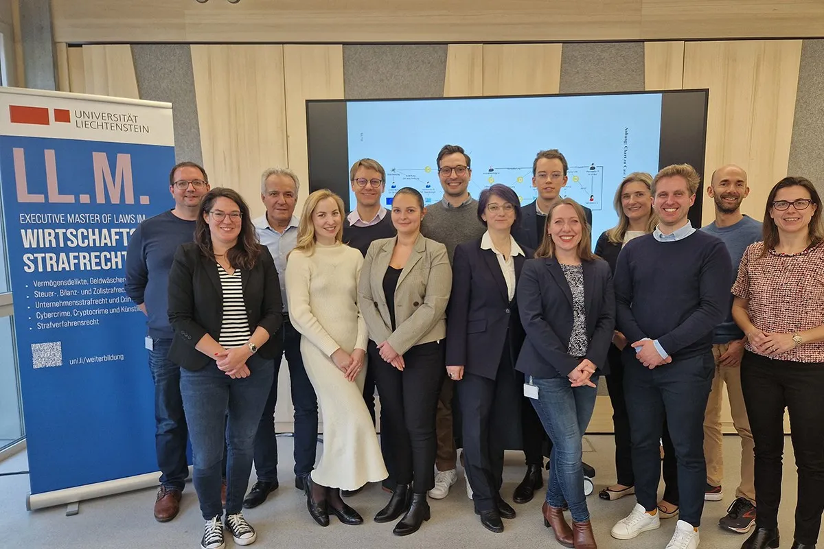 Gruppenfoto von Studierenden und Dozenten des Executive Masters of Law im Wirtschaftsstrafrecht an der Universität Liechtenstein. Im Hintergrund ein Bildschirm mit Präsentationsfolie und ein Roll-up mit Informationen zum Studiengang.