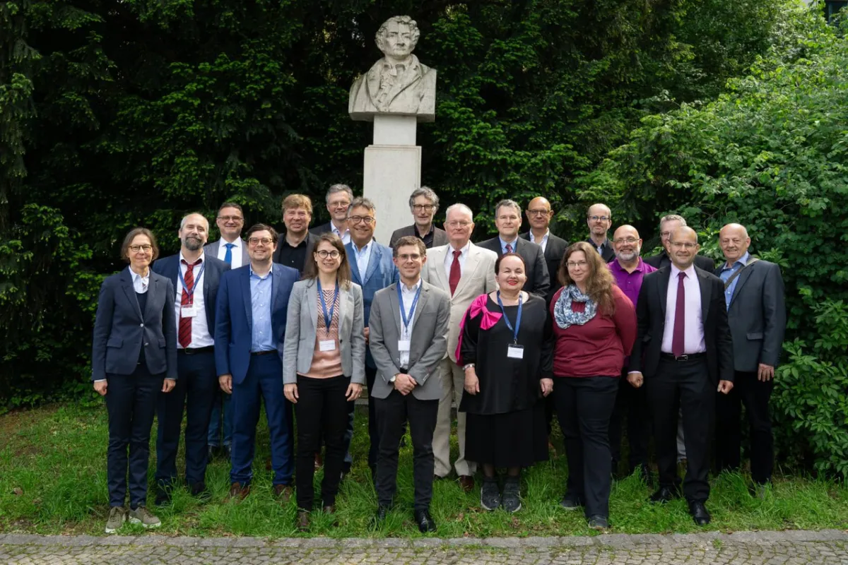 Eine Gruppe von Menschen steht vor einer Büste von Paul Johann Anselm von Feuerbach im Freien. Die Personen tragen formelle Kleidung und lächeln in die Kamera. Im Hintergrund sind grüne Büsche und Bäume zu sehen.