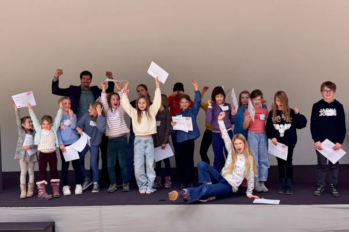 Eine Gruppe von Kindern und einem Erwachsenen steht auf einer Bühne und hält stolz Zertifikate hoch. Die Kinder wirken fröhlich und begeistert. Sie nehmen an einem Fundraising-Workshop an der Universität Liechtenstein teil.