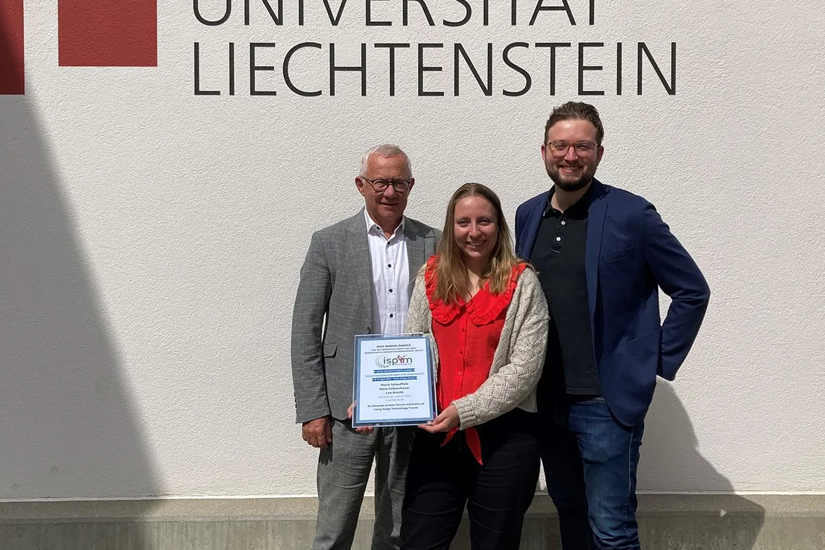 Drei Personen stehen vor einer Wand mit dem Logo der Universität Liechtenstein. Eine Person hält eine Auszeichnung in den Händen.