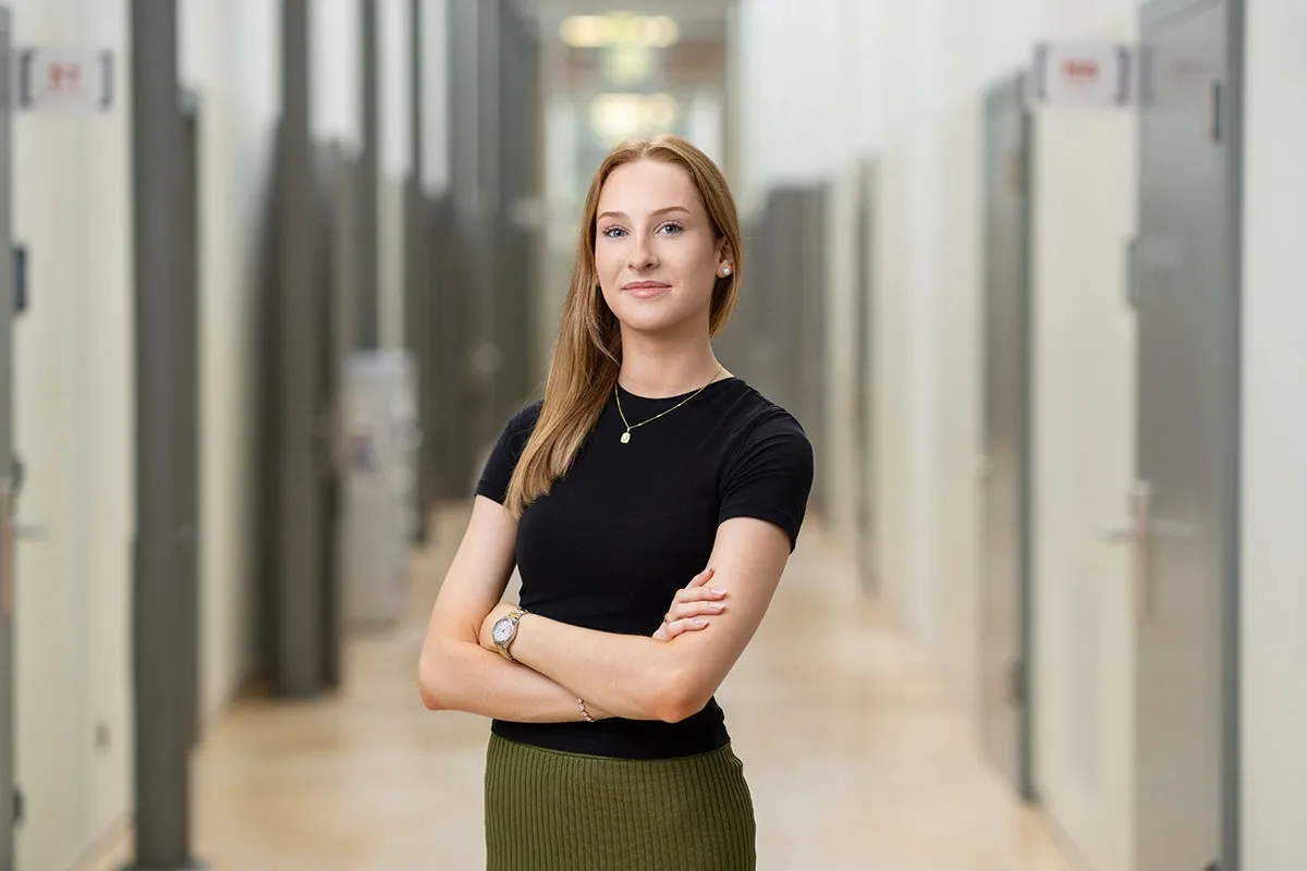 Eine junge Frau steht selbstbewusst mit verschränkten Armen in einem modernen Flur. Sie trägt ein schwarzes Oberteil und einen grünen Rock.