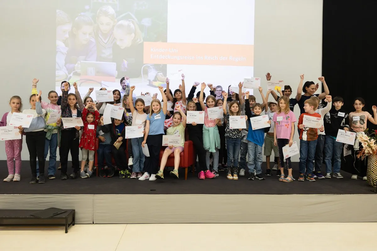 Gruppe von Kindern auf einer Bühne, die stolz Zertifikate hochhalten, im Rahmen der Kinder-Uni an der Universität Liechtenstein. Im Hintergrund ein Bildschirm mit der Aufschrift „Entdeckungsreise ins Reich der Regeln“.