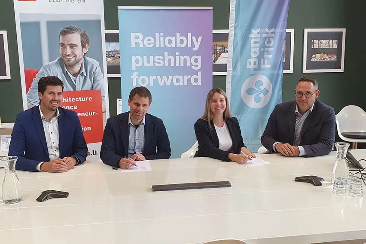 Vier Personen sitzen an einem Konferenztisch und unterzeichnen Dokumente. Im Hintergrund sind Banner der Universität Liechtenstein und Bank Frick zu sehen. Die Szene symbolisiert die Verlängerung der Kooperation zwischen der Universität und der Bank.