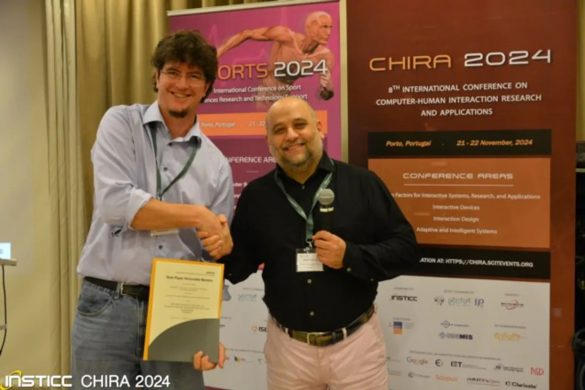 Zwei Männer stehen vor Bannern der CHIRA 2024 Konferenz. Einer hält ein Zertifikat, während der andere ein Mikrofon hält. Beide lächeln und schütteln sich die Hand. Im Hintergrund sind Details zur Konferenz in Porto, Portugal, sichtbar.