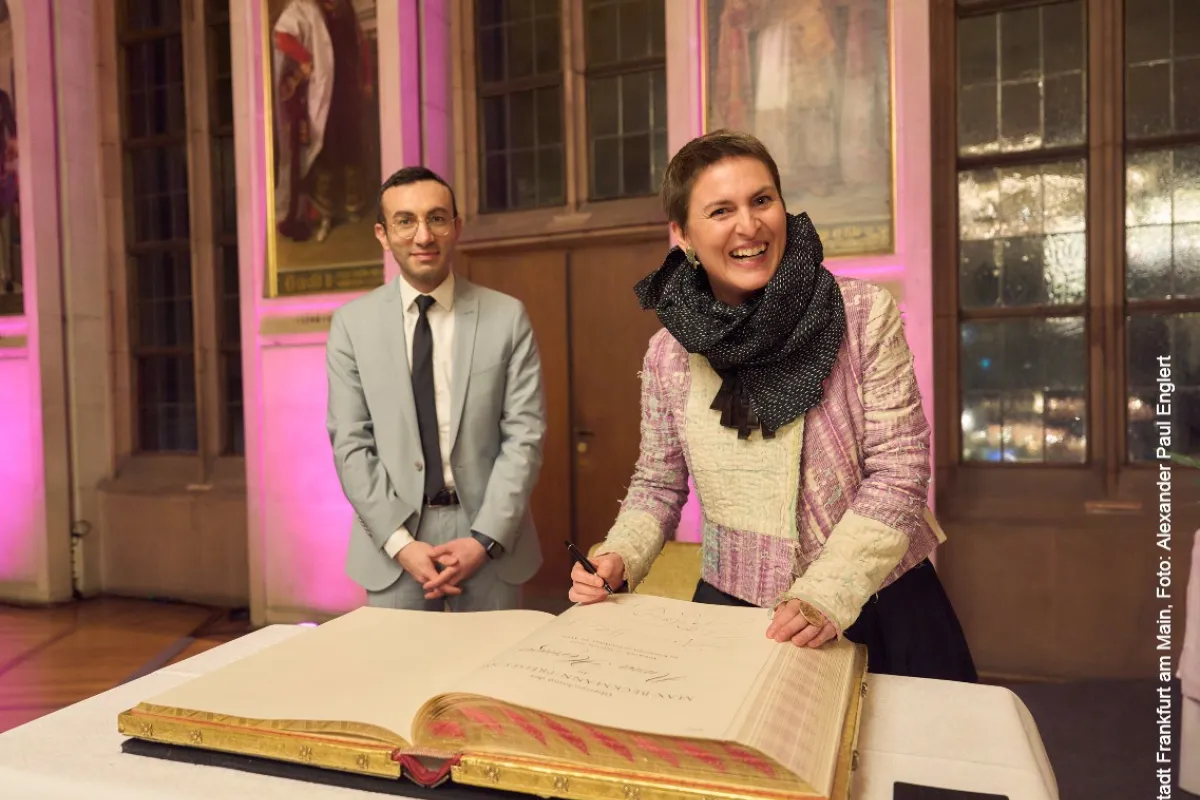 Eine Person in einem eleganten Outfit unterschreibt ein großes Buch auf einem Tisch, während eine andere Person daneben steht. Der Raum ist festlich beleuchtet und im Hintergrund sind Gemälde an der Wand zu sehen.