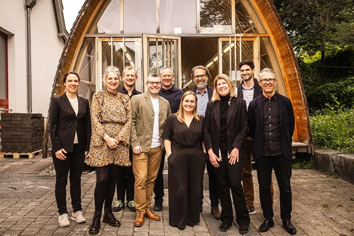 Eine Gruppe von zehn Personen steht lächelnd vor einem modernen Holzgebäude mit großen Fenstern. Die Szene vermittelt eine freundliche und professionelle Atmosphäre.