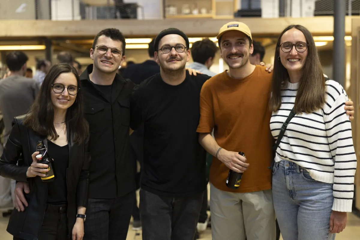 Eine Gruppe von fünf Personen lächelt in die Kamera bei einem Indoor-Event. Zwei Personen halten Getränke in der Hand. Die Umgebung ist lebhaft und gut beleuchtet, was auf eine gesellige Atmosphäre hinweist.