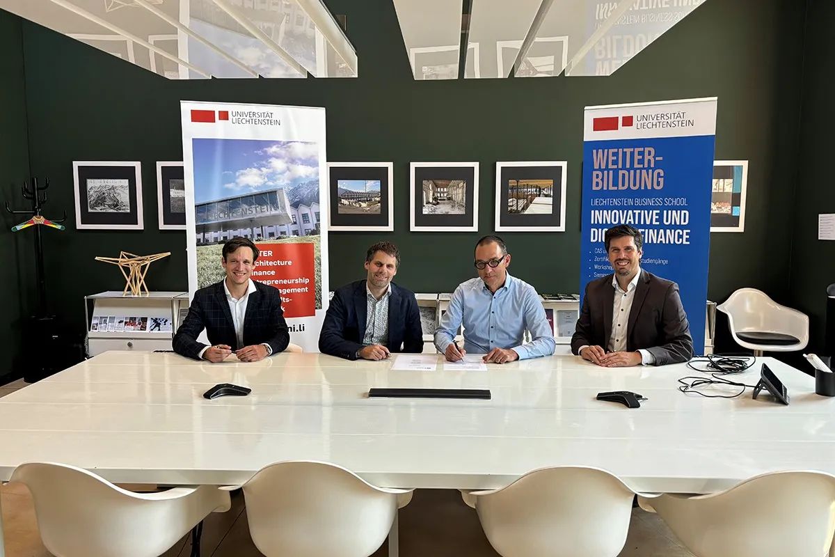 Vier Männer sitzen an einem Tisch in einem modernen Konferenzraum der Universität Liechtenstein. Im Hintergrund sind Banner mit dem Logo der Universität und Informationen zur Weiterbildung sichtbar.