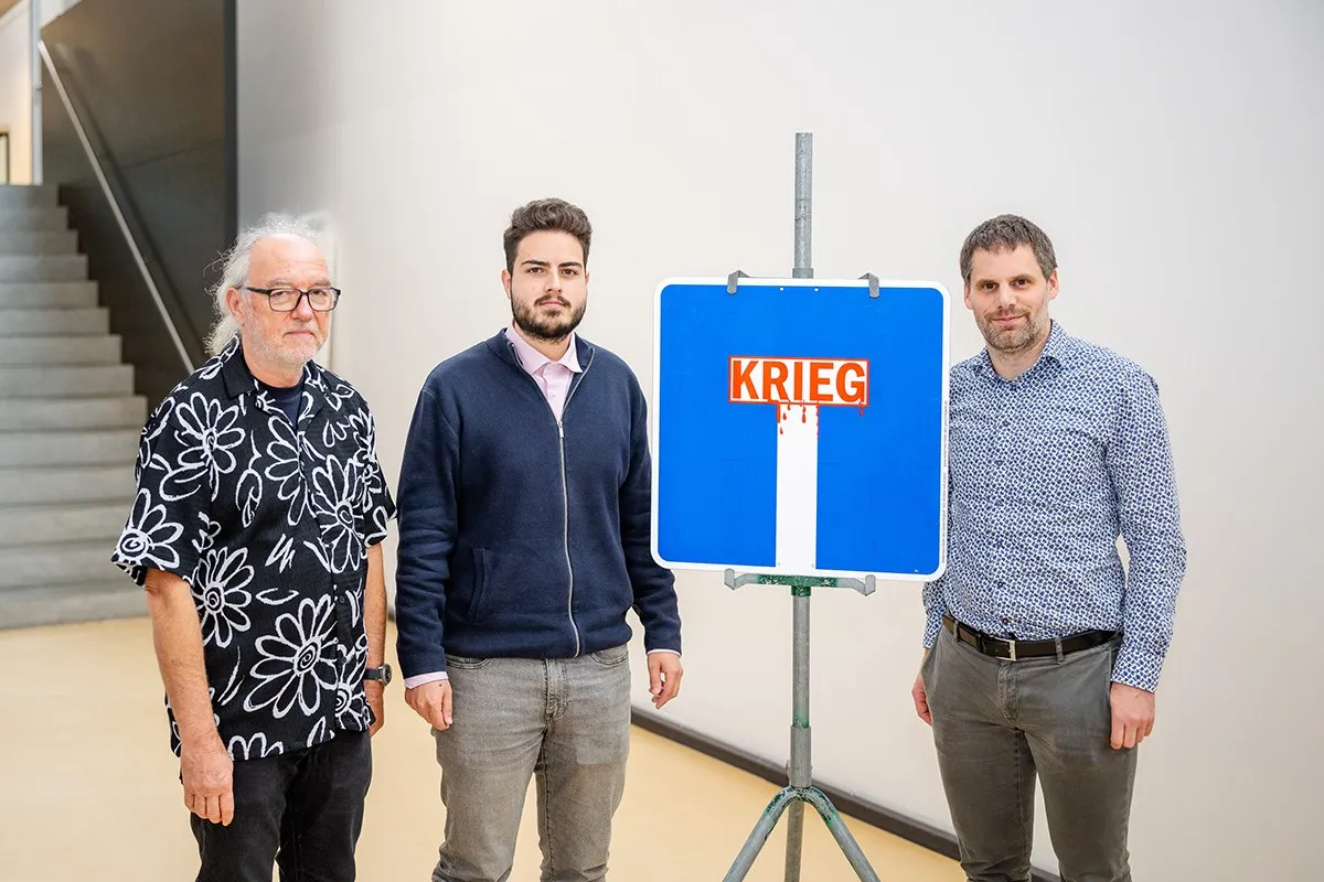 Drei Männer stehen neben einem blauen Verkehrsschild mit der Aufschrift "Krieg", das symbolisch eine Sackgasse darstellt. Das Bild wurde bei der Veranstaltung "Verkehrsschilder der Gerechtigkeit" an der Universität Liechtenstein aufgenommen.