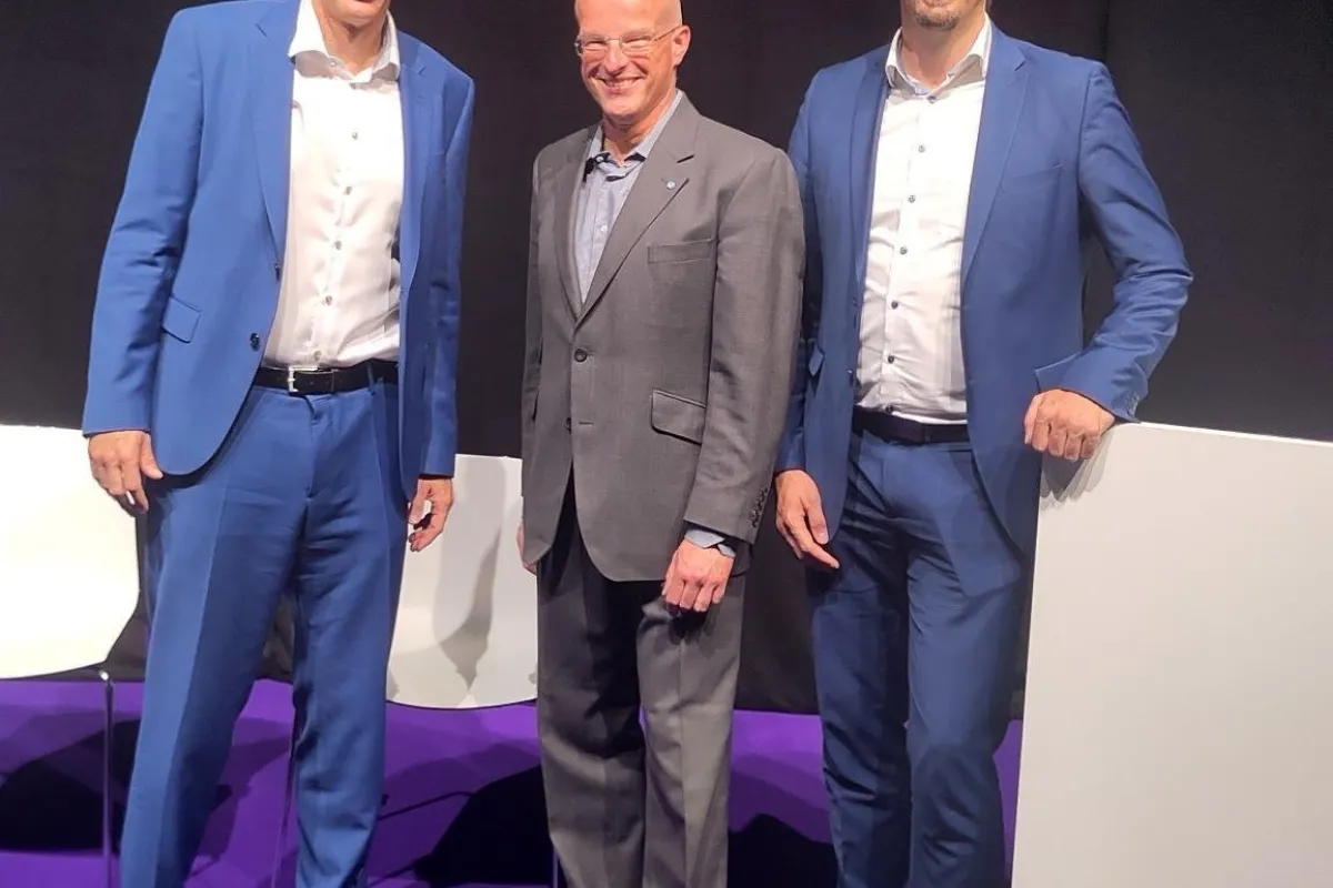 Drei Männer in Anzügen stehen auf einer Bühne mit lila Teppich. Sie lächeln in die Kamera. Im Hintergrund sind schwarze Vorhänge zu sehen.