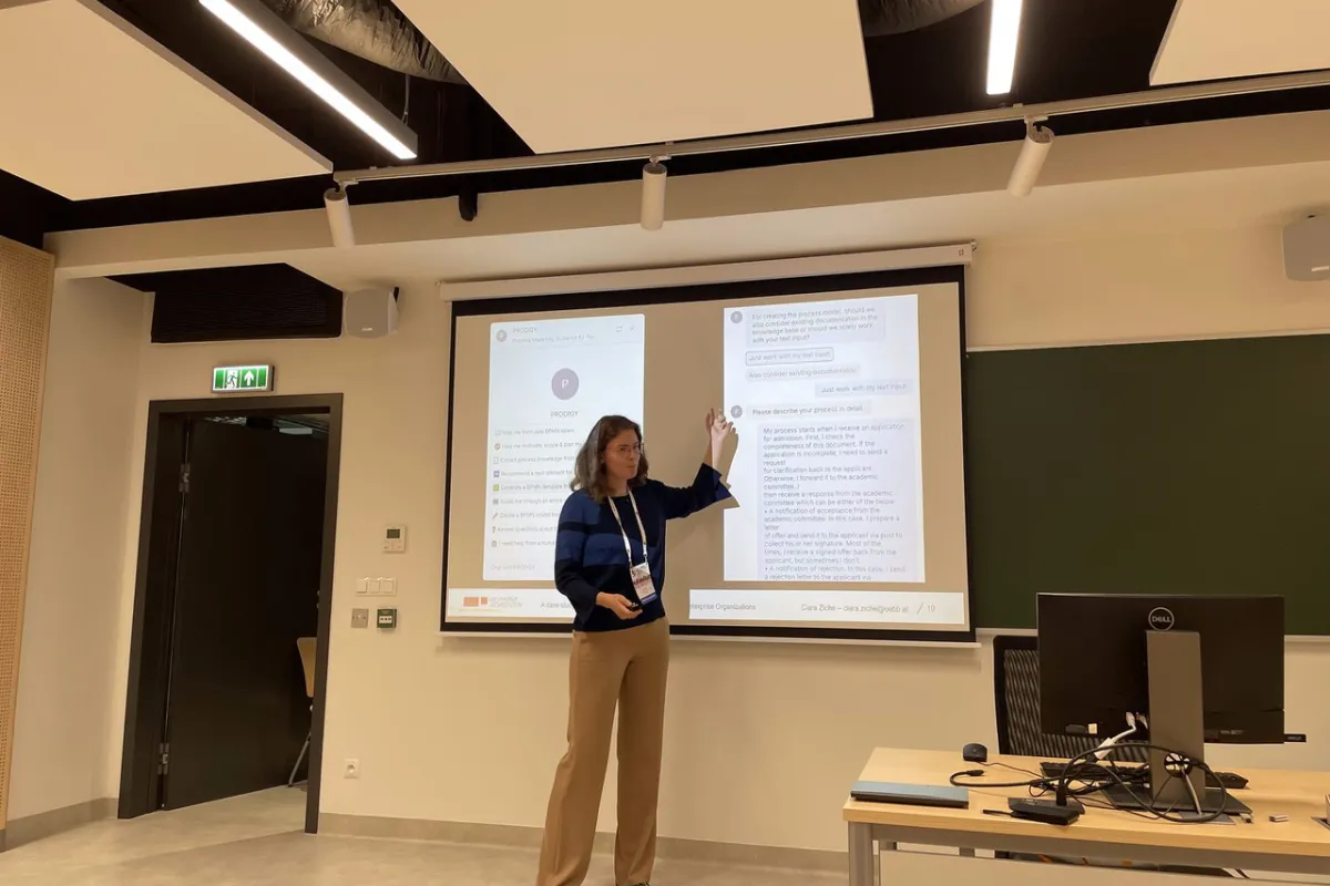 Eine Person präsentiert in einem modernen Seminarraum vor einer Leinwand mit zwei projizierten Dokumenten. Im Vordergrund steht ein Tisch mit einem Computerbildschirm.