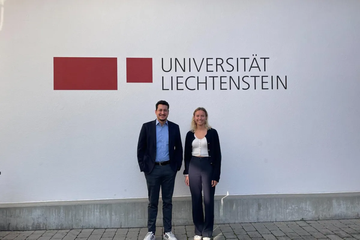 Zwei Personen stehen vor einer Wand mit dem Logo der Universität Liechtenstein. Sie lächeln in die Kamera.