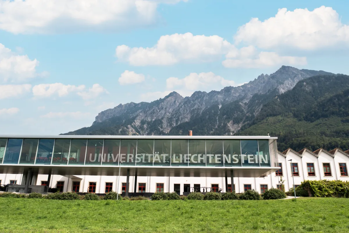 Glasfassade der Universität Liechtenstein vor Bergkulisse bei blauem Himmel.