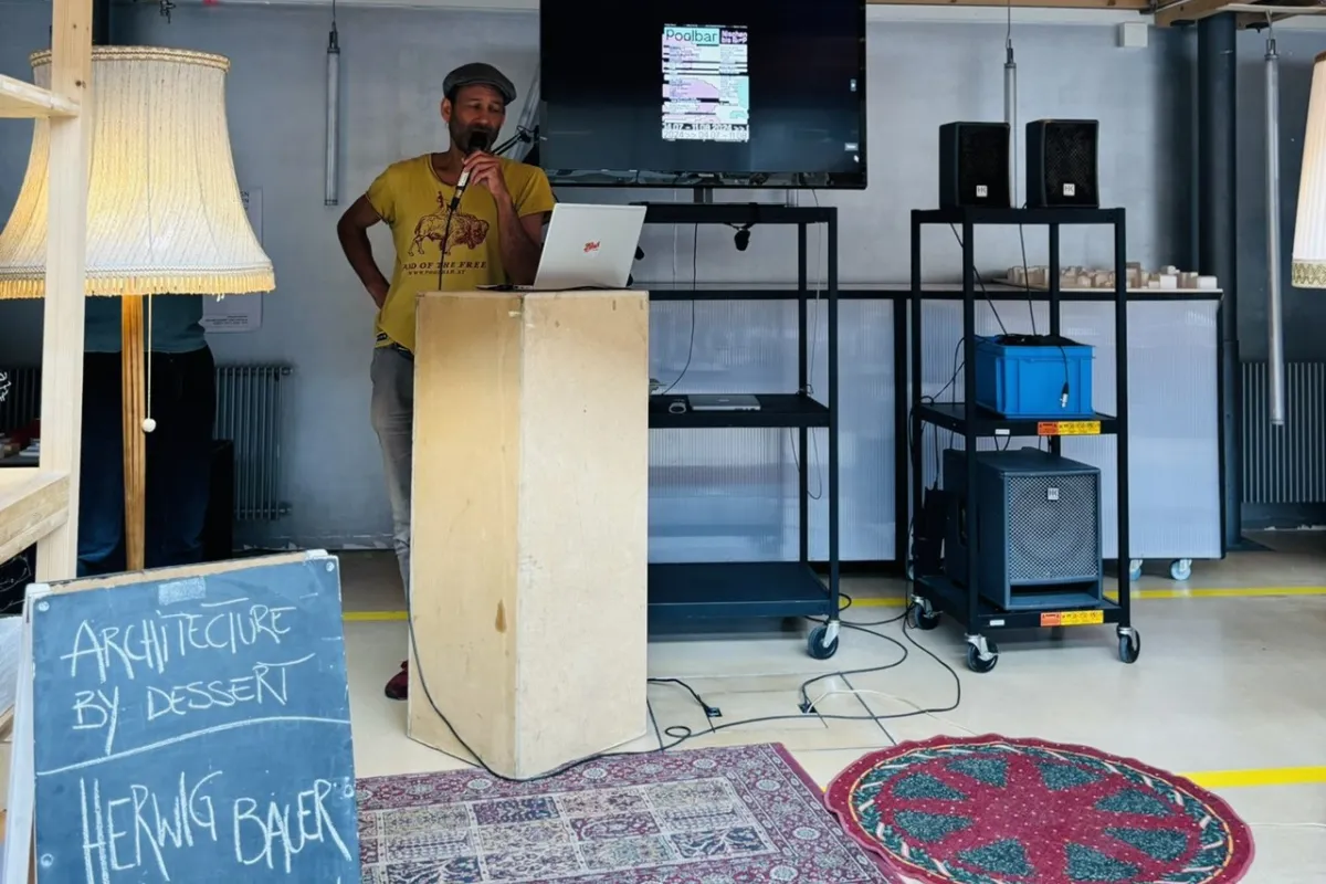 Ein Mann steht an einem Rednerpult in einem Raum mit Holzdach. Neben ihm ist ein Bildschirm mit einer Präsentation und ein Schild mit der Aufschrift „Architecture by Dessert, Herwig Bauer, Poolbarfestival“. Der Raum ist mit Teppichen und Stehlampen dekoriert.