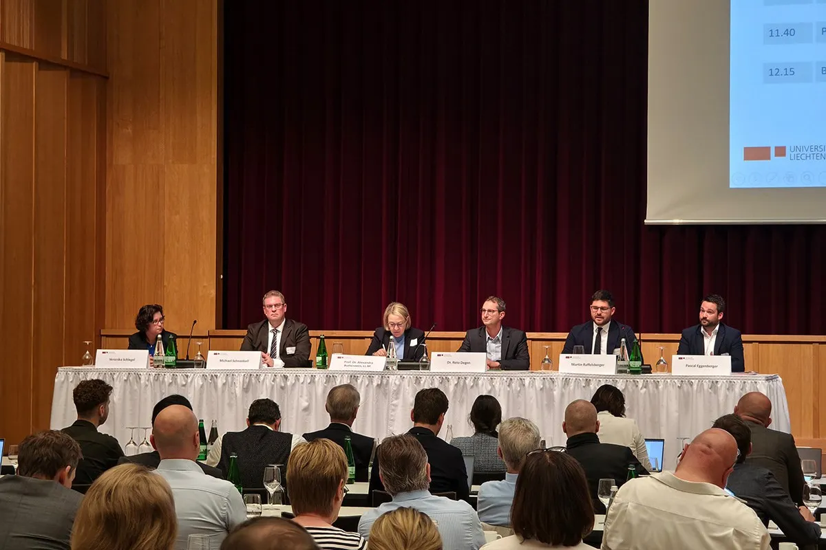 Ein Podium mit sechs Personen sitzt vor einem Publikum in einem Konferenzsaal. Auf dem Tisch stehen Namensschilder und Wasserflaschen. Im Hintergrund ist ein roter Vorhang und ein Teil einer Präsentationsleinwand zu sehen.