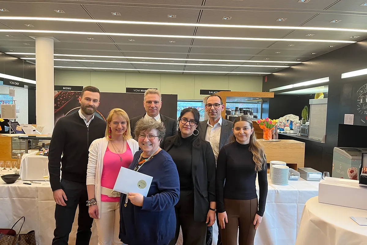 Eine Gruppe von sieben Personen steht lächelnd in einem modernen Raum. Eine Person hält ein Zertifikat in den Händen. Im Hintergrund sind Tische mit Geschirr und Dekorationen zu sehen.
