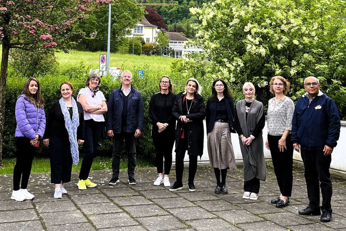 Eine Gruppe von zehn Personen steht draußen auf einem gepflasterten Weg vor einer grünen Hecke und blühenden Bäumen. Die Personen sind in Freizeitkleidung und lächeln in die Kamera. Im Hintergrund sind ein Gebäude und eine Wiese zu sehen.