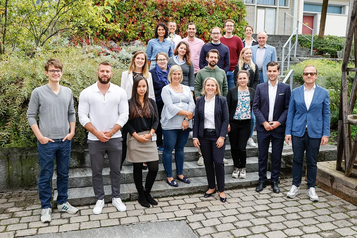 Eine Gruppe von Studierenden und Dozenten des Executive Master of Laws (LL.M.) im Gesellschafts-, Stiftungs- und Trustrecht steht auf einer Treppe im Freien. Sie lächeln in die Kamera, umgeben von grüner Vegetation und einem Gebäude im Hintergrund.