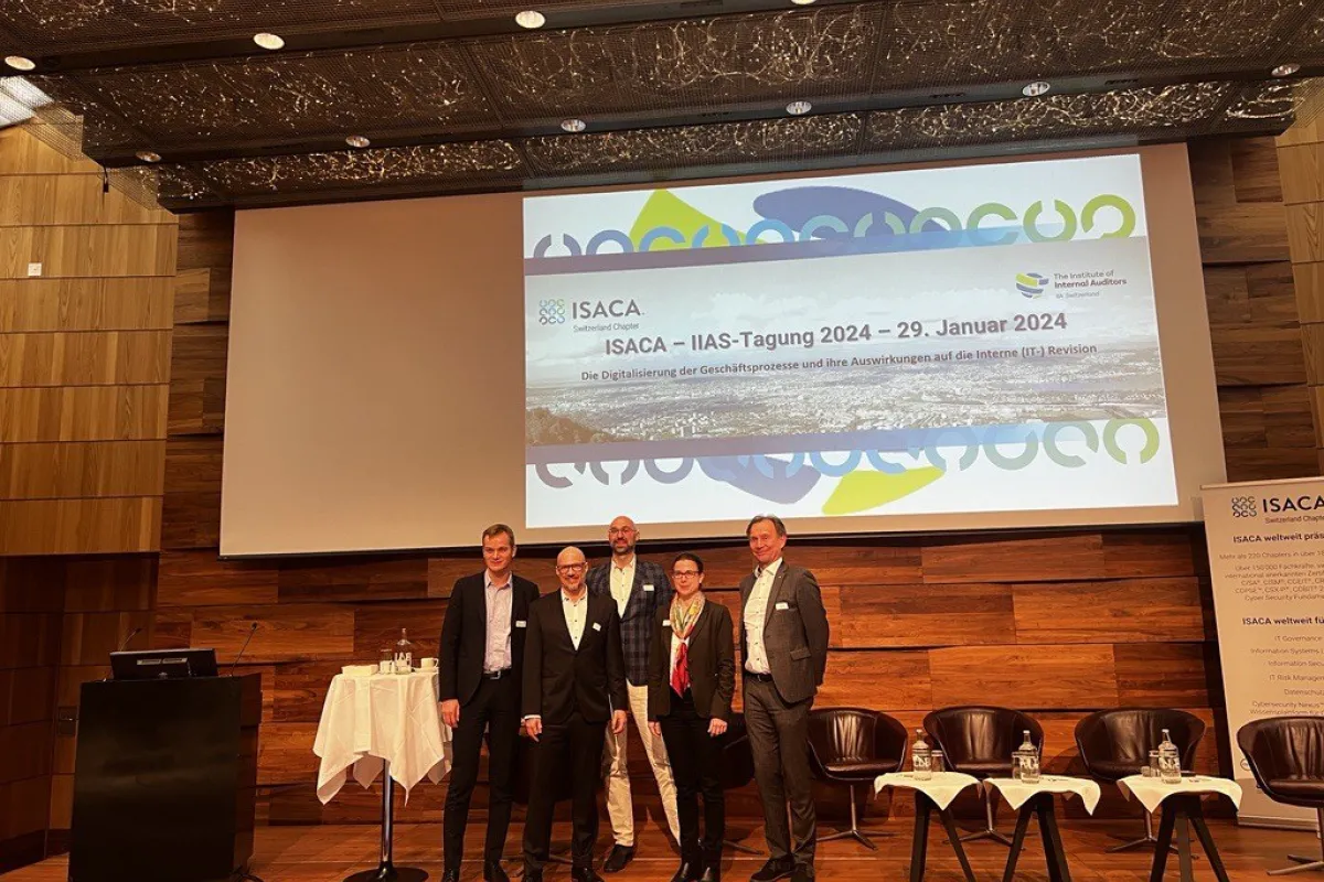 Gruppe von fünf Personen steht auf einer Bühne vor einer Leinwand mit dem Titel der ISACA-IIAS-Tagung 2024 in Zürich. Im Hintergrund sind Stühle und ein Tisch mit Wasserflaschen zu sehen.