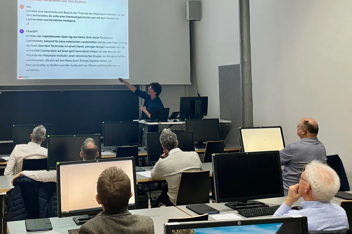 Ein Dozent hält einen interaktiven Workshop an der Universität Liechtenstein. Er zeigt auf eine Präsentation über künstliche Intelligenz, während die Teilnehmenden an Computern sitzen und aufmerksam zuhören.