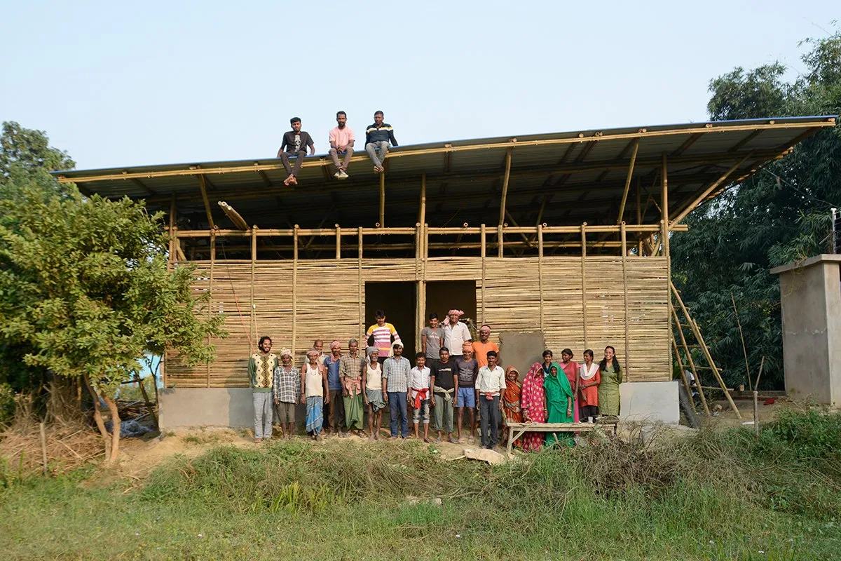 Eine Gruppe von Menschen steht vor einem im Bau befindlichen Gebäude aus Bambus in Bahuarwa, Indien. Drei Personen sitzen auf dem Dach. Das Bild zeigt den Fortschritt eines Schulbauprojekts, das von der Universität Liechtenstein unterstützt wird.