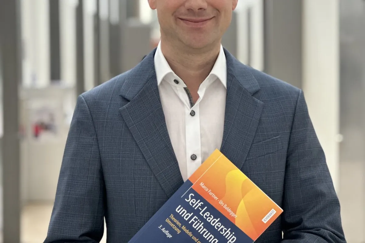 Ein Mann in einem blauen Anzug hält das Buch „Self-Leadership und Führung“ in einem modernen, hellen Flur.