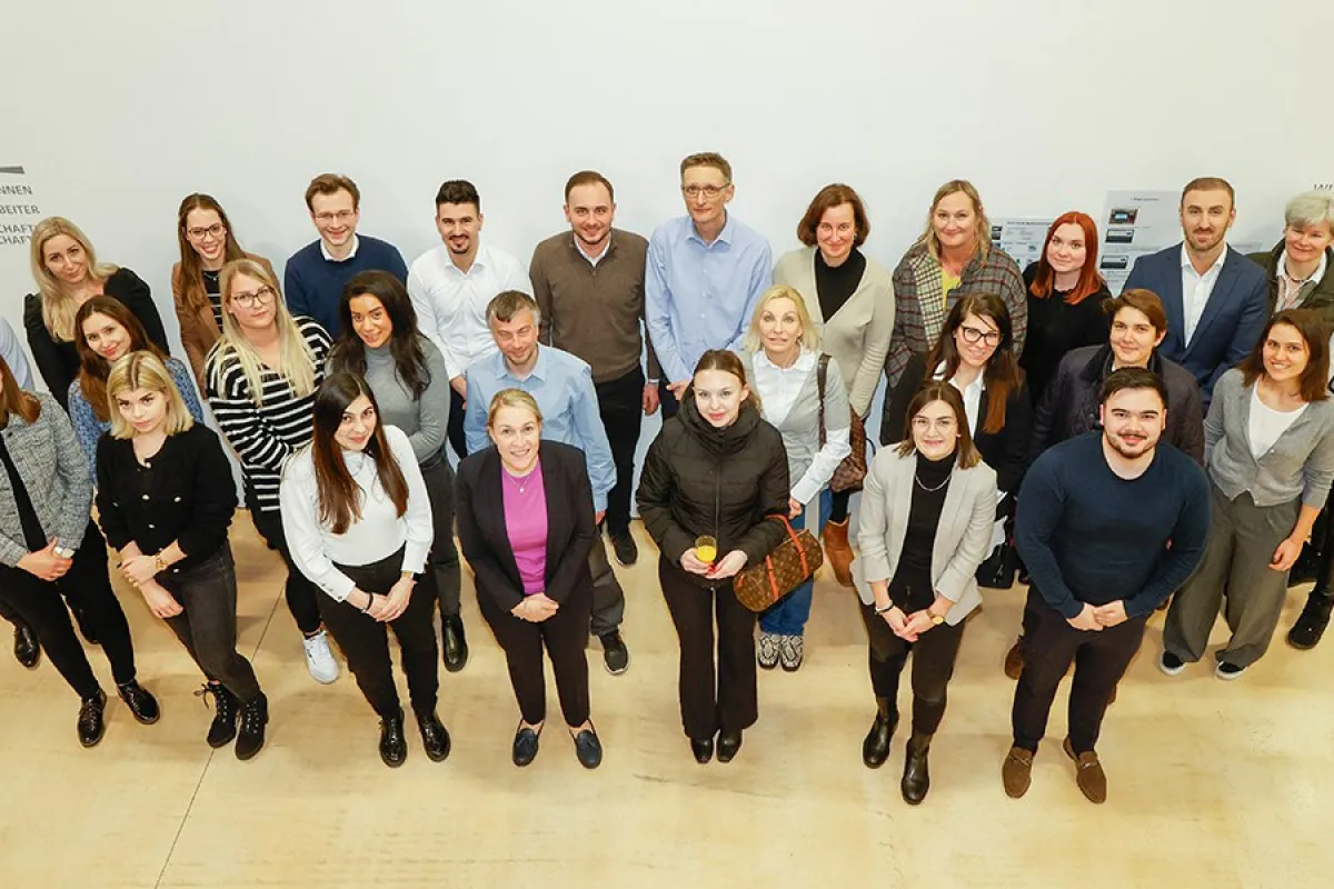 Teilnehmende des Zertifikatsstudiengangs Treuhandwesen an der Universität Liechtenstein stehen in einer Gruppe zusammen und blicken nach oben. Sie sind in einem hellen Raum mit neutralem Hintergrund.