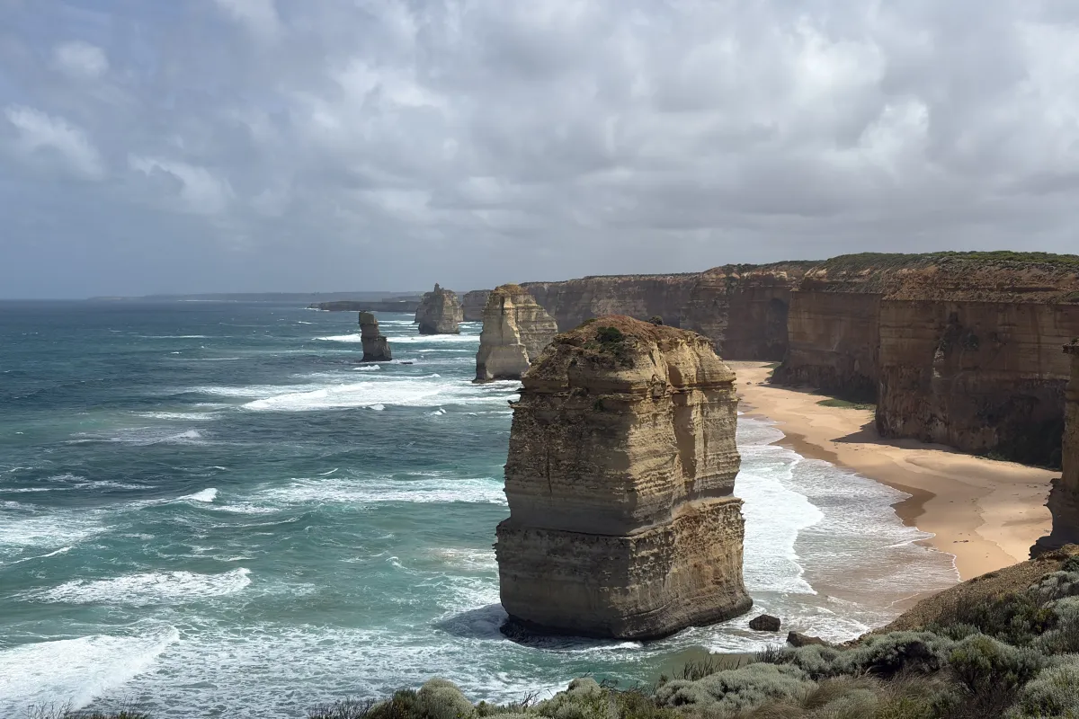 Steile Felsklippen und markante Felsnadeln ragen aus dem Meer entlang einer wilden Küste mit Sandstrand und brechenden Wellen.