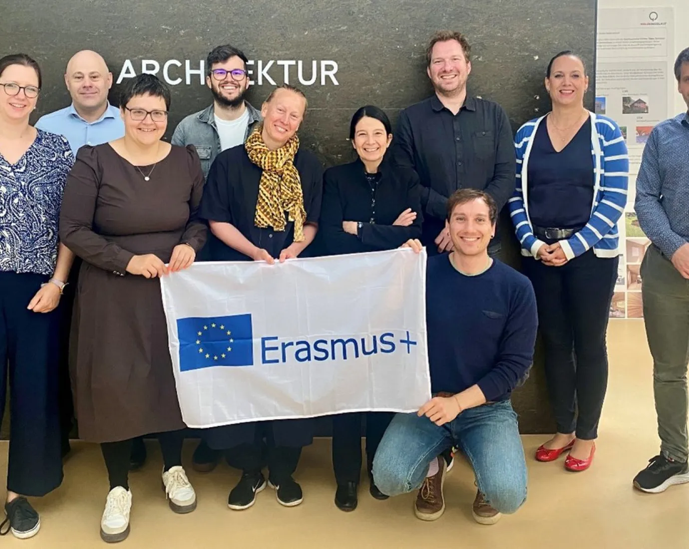 Gruppe von zehn Personen steht vor einer Wand mit dem Schriftzug "Architektur" und hält ein Erasmus+ Banner. Sie lächeln in die Kamera.