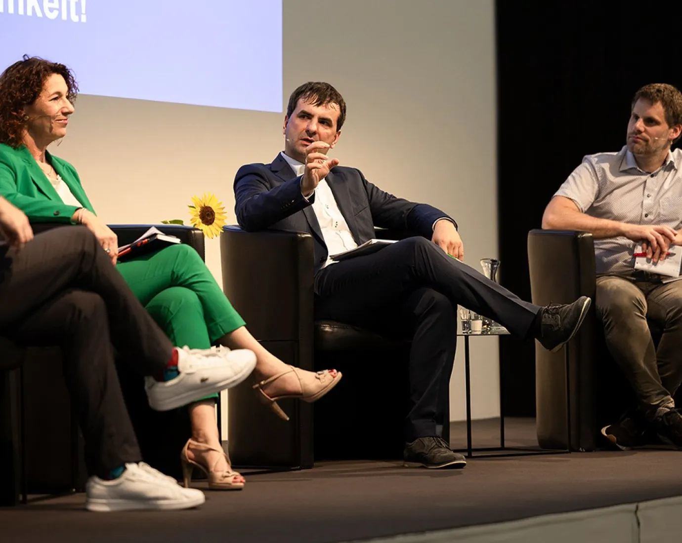 Podiumsdiskussion an der Universität Liechtenstein: Drei Personen sitzen auf einer Bühne in schwarzen Sesseln und diskutieren angeregt. Eine Frau in grünem Anzug und zwei Männer in formeller Kleidung sind zu sehen. Im Hintergrund ist ein Teil einer Leinwand mit Text sichtbar.