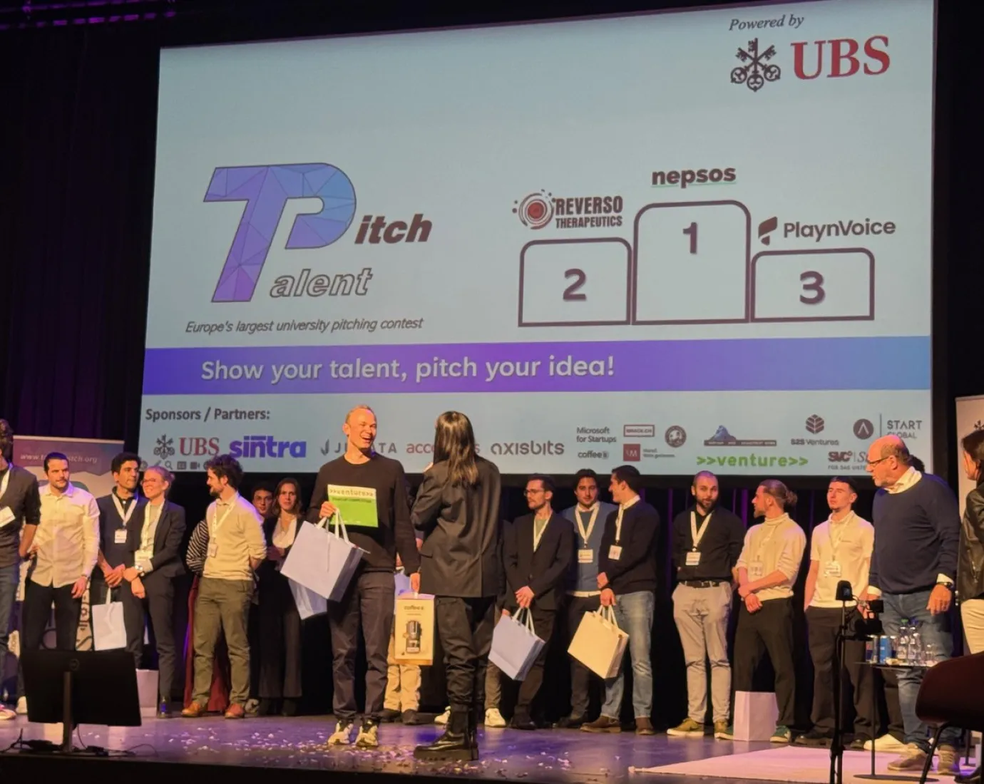 Eine Gruppe von Menschen steht auf einer Bühne beim Talent Pitch Switzerland im Volkshaus Zürich. Im Hintergrund ist eine Leinwand mit den Logos der Gewinner-Start-ups Reverso Therapeutics, Nepsos und PlaynVoice zu sehen. Die Veranstaltung wird von UBS gesponsert.