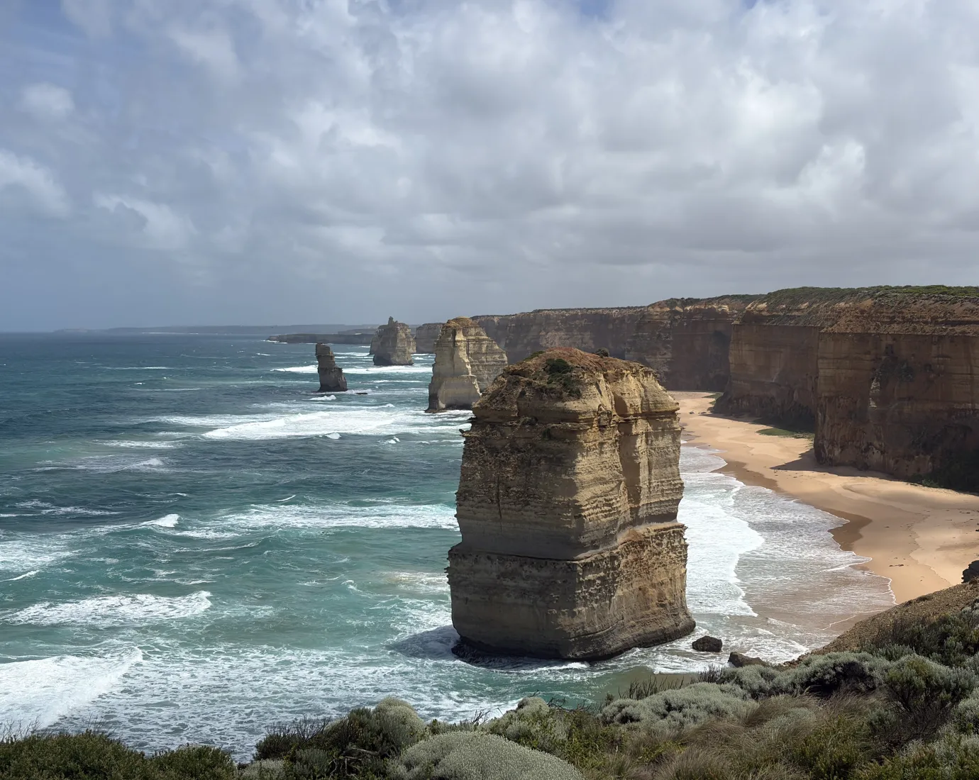 Steile Felsklippen und markante Felsnadeln ragen aus dem Meer entlang einer wilden Küste mit Sandstrand und brechenden Wellen.