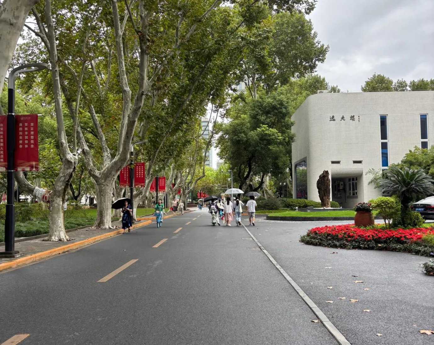 Baumgesäumte Allee auf einem Campus mit Spaziergängern, roten Bannern und einem modernen Gebäude am Straßenrand.