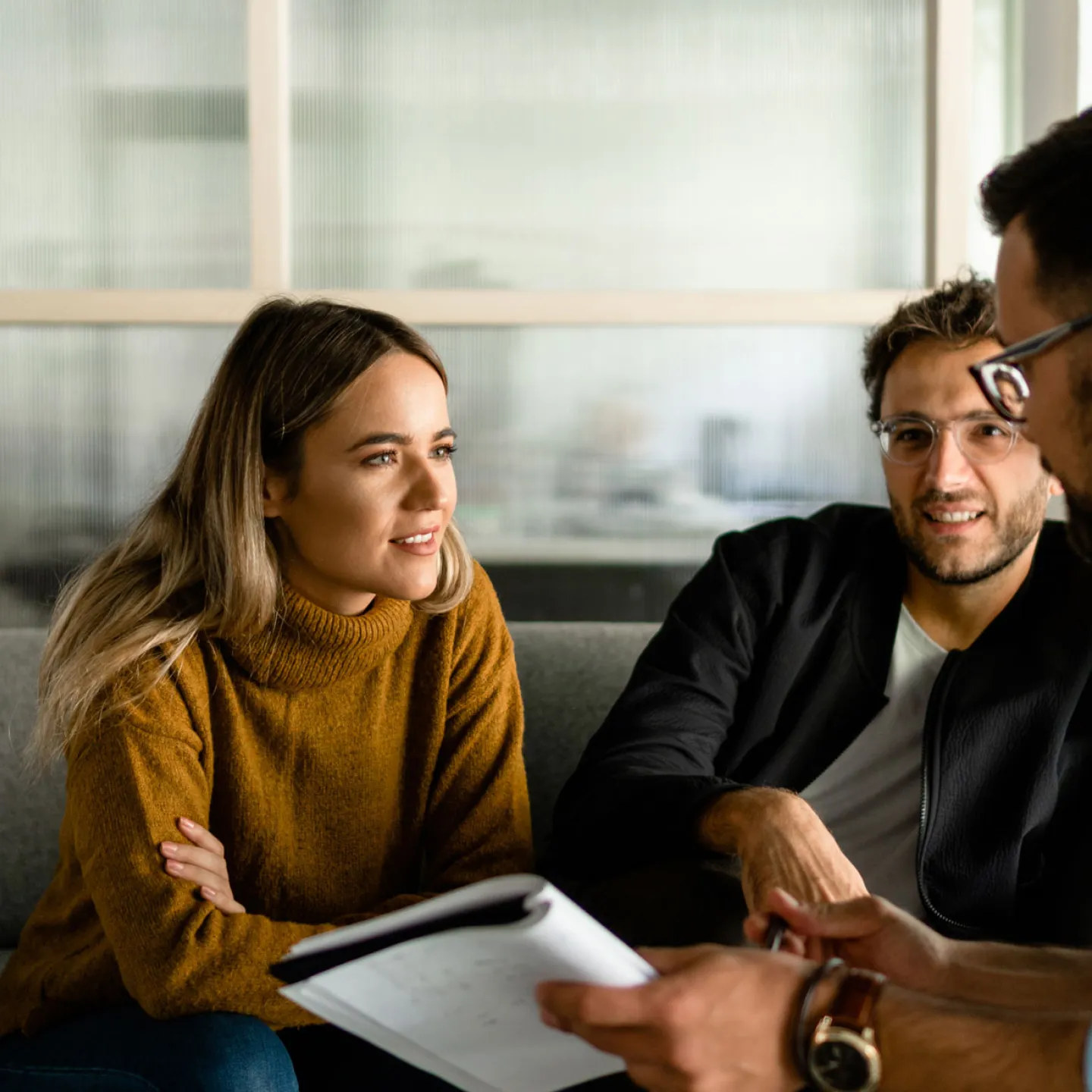 Eine junge Frau und ein junger Mann sitzen nebeneinander auf einem Sofa, während der dritte, der ihnen gegenüber sitzt, sich mit einem aufgeschlagenen Buch in der Hand nach vorne beugt. Der dritte Person führt ein angeregtes Gespräch mit den beiden über das Thema Verantwortung und Gesellschaft. Die konzentrierten Gesichtsausdrücke und die gestikulierende Körpersprache der Gesprächsteilnehmer unterstreichen die Intensität der Diskussion.