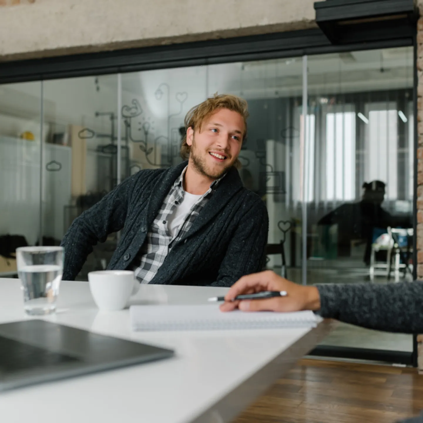 Zwei Männer in einem modernen Büro unterhalten sich an einem Tisch. Der Mann im Vordergrund schreibt in ein Notizbuch, während der andere Mann, der zurückgelehnt und lächelnd ist, zuhört. Auf dem Tisch stehen ein Laptop, ein Glas Wasser und eine Tasse. Im Hintergrund sind Glaswände und Ziegelwände zu sehen, die dem Raum eine offene und zeitgemäße Atmosphäre verleihen.