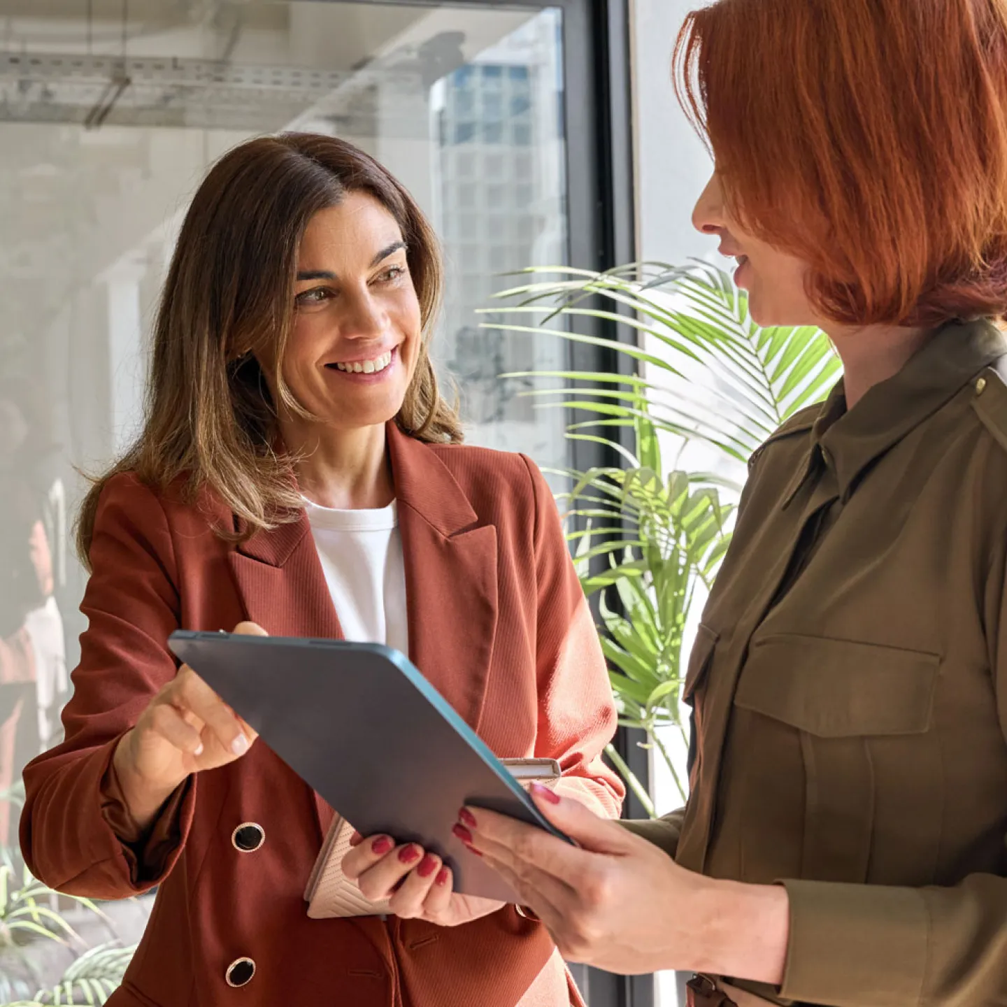 Zwei Frauen stehen in einem modernen Bürogebäude und unterhalten sich. Beide sind formell gekleidet und wirken konzentriert. Im Hintergrund sind große Fenster zu sehen, die viel natürliches Licht in den Raum lassen, sowie Büromöbel und Pflanzen, die eine professionelle Atmosphäre schaffen.
