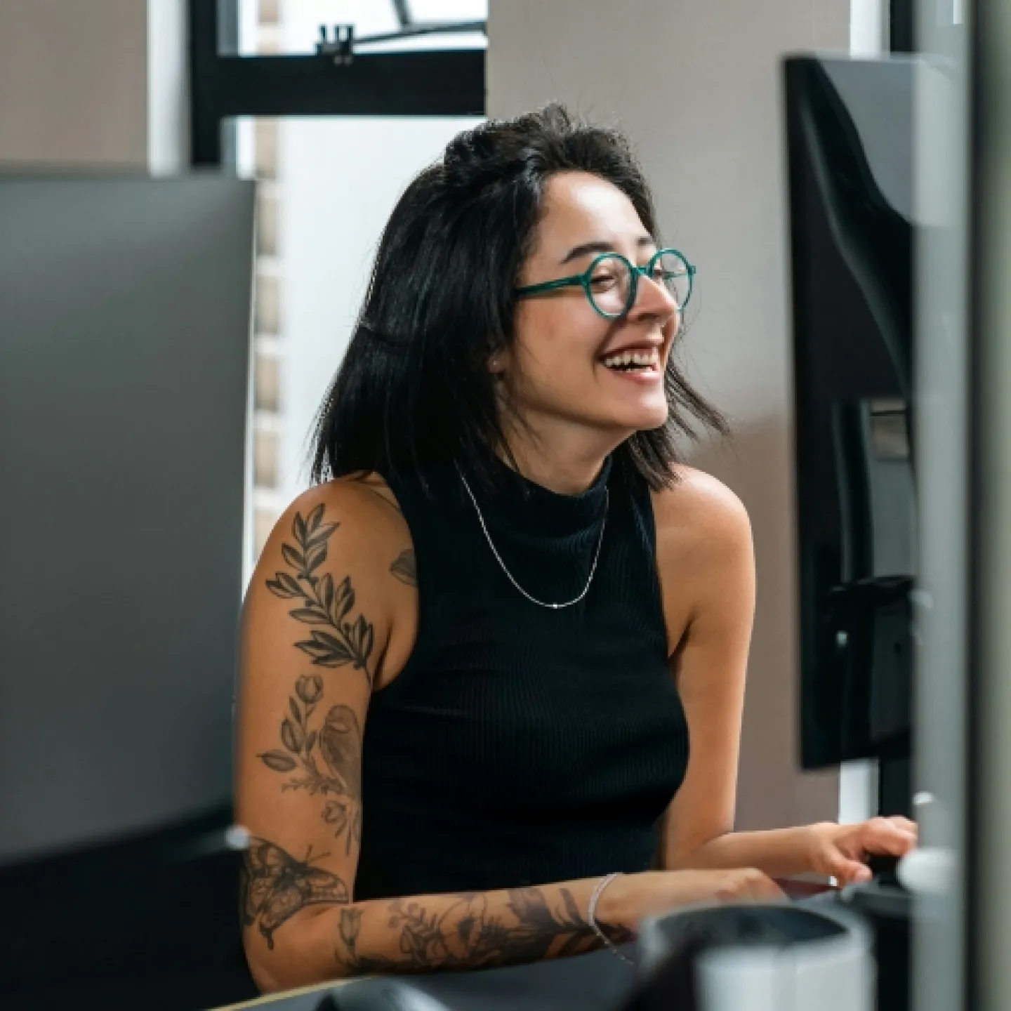 Eine junge lächelnde Frau, mit schulterlangem schwarzen Haar und Brille, in ärmellosem Shirt, das zahlreiche Tattoos entblößt. Sie sitzt vor einem Computer.