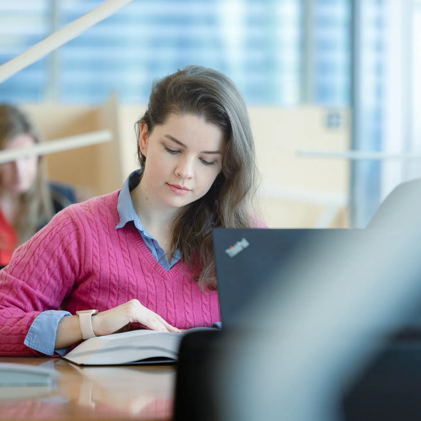 Studentin der Universität Liechtenstein arbeitet konzentriert mit Buch und Laptop in der lichtdurchfluteten Bibliothek.