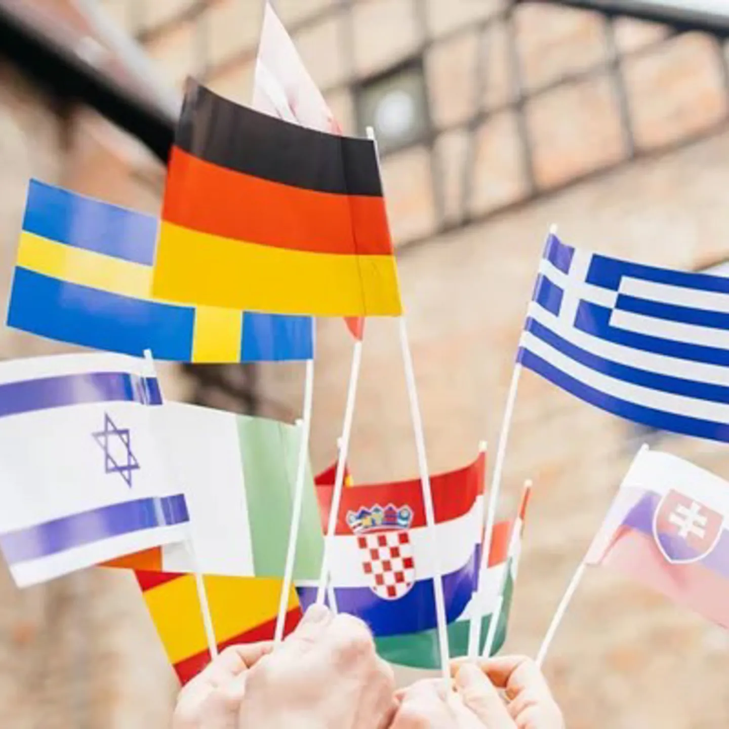 Mehrere Hände halten kleine internationale Flaggen in die Höhe, darunter unter anderem die Flaggen von Deutschland, Griechenland, Schweden, Israel, Kroatien, der Slowakei, Italien und Litauen. Im Hintergrund ist ein historisches Gebäude zu sehen. Das Bild symbolisiert internationale Vielfalt und kulturellen Austausch an der Universität Liechtenstein.