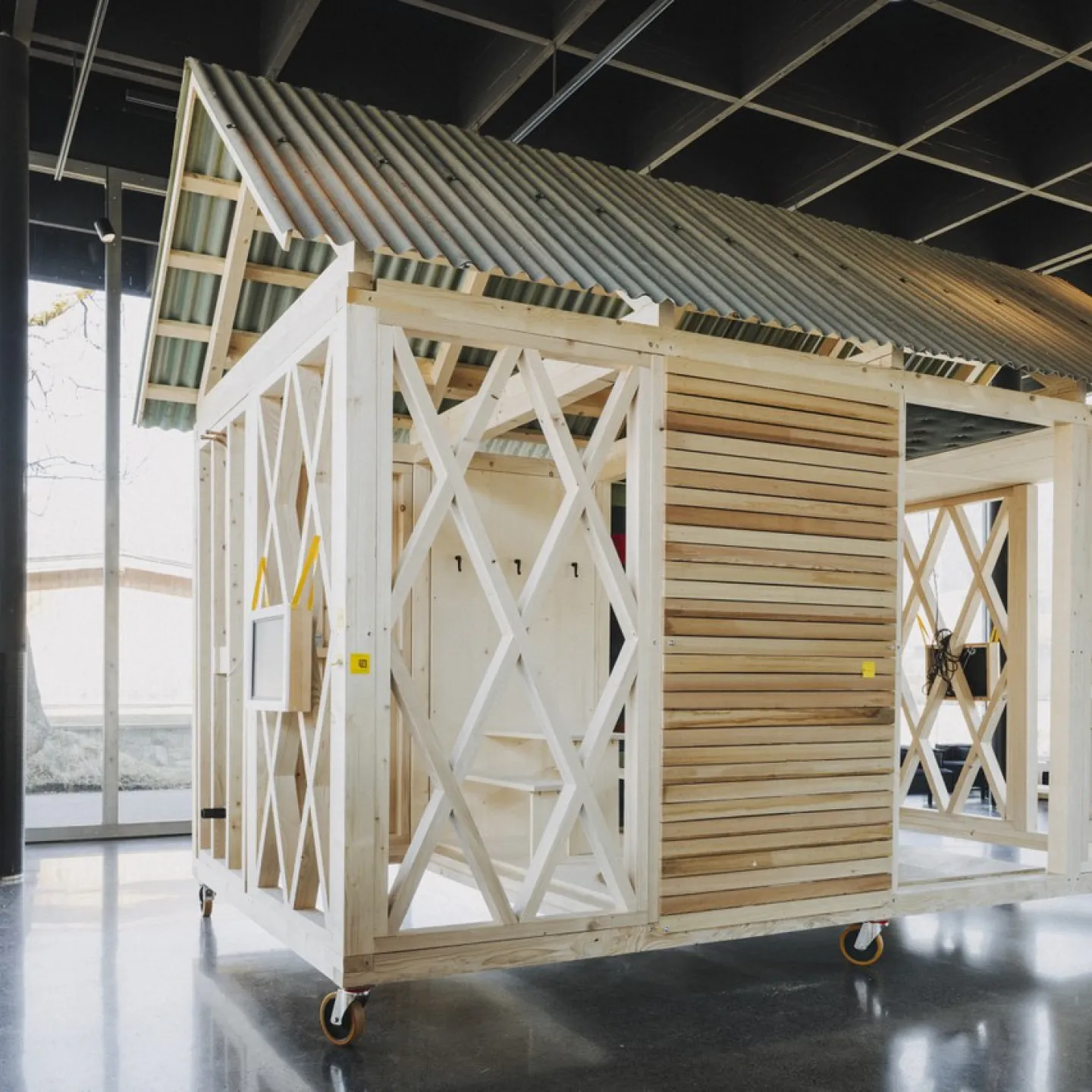 Ein einfaches, modulares Holzgebäude mit einem Wellblechdach steht auf Rollen in einem modernen, lichtdurchfluteten Raum. Das Design zeigt eine offene Struktur mit diagonalen Verstrebungen und Holzverkleidung, ideal für nachhaltiges Bauen und architektonische Bildung.