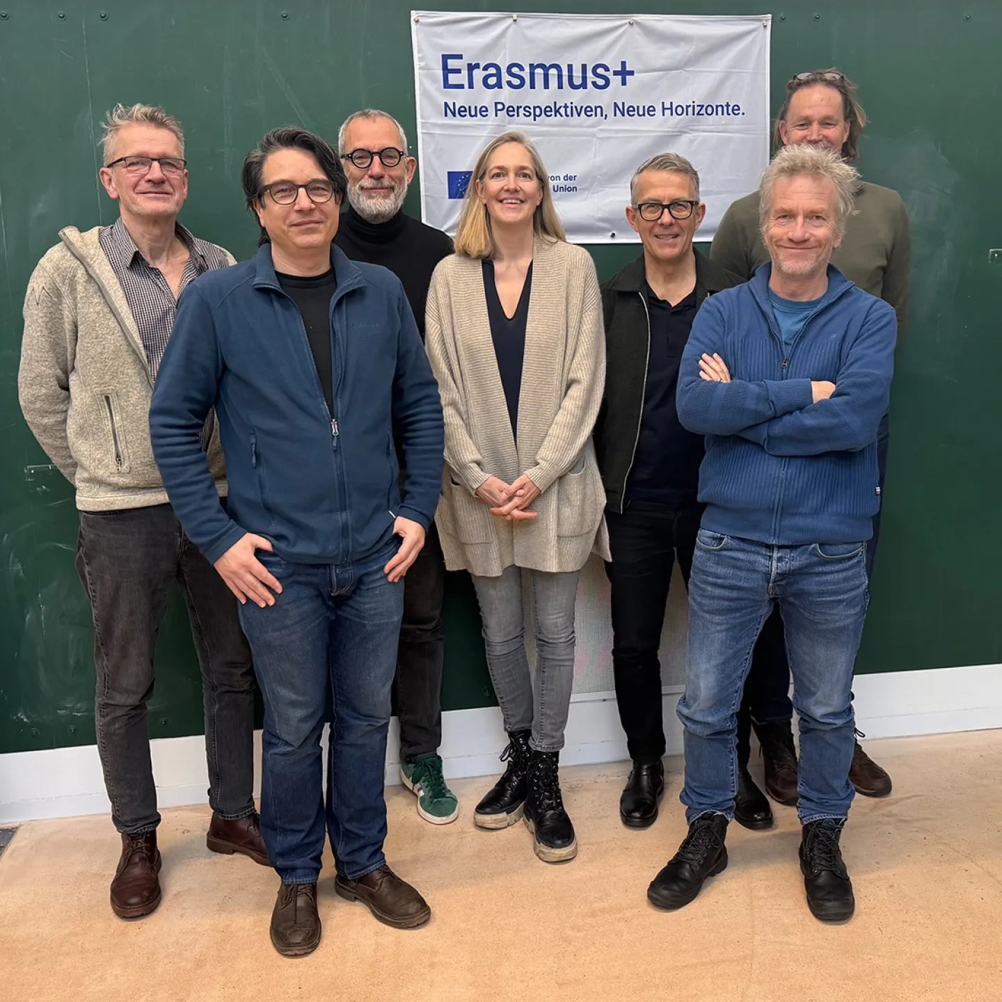 Eine Gruppe von acht Personen steht vor einem grünen Hintergrund. Sie lächeln in die Kamera. Im Hintergrund hängt ein Banner mit der Aufschrift "Erasmus+ Neue Perspektiven, Neue Horizonte".